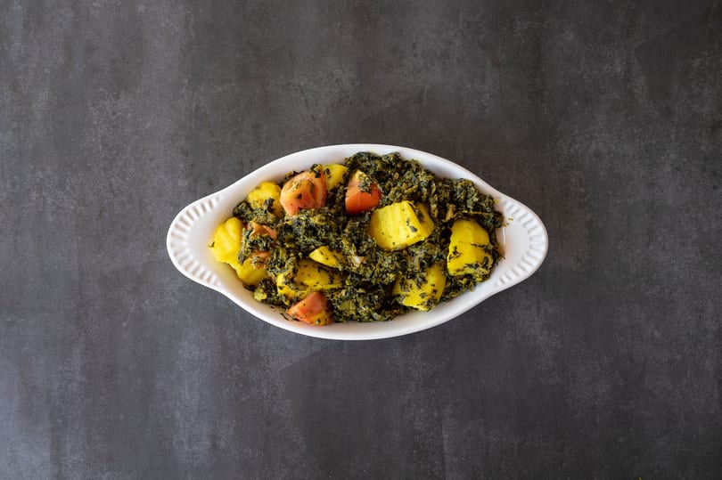 A white oval dish filled with saag aloo, a green spinach and potato curry with tomatoes.