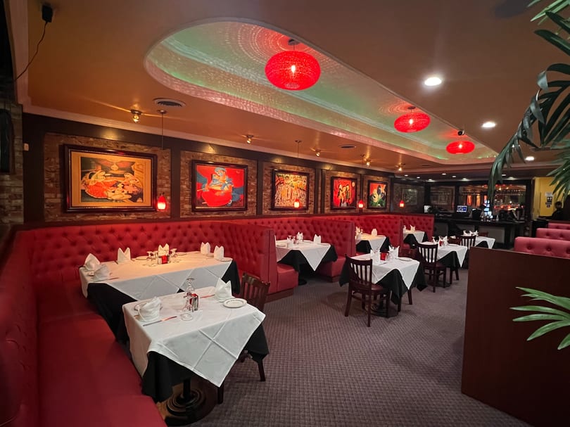 Restaurant interior with red booth seating, white-clothed tables, and colorful framed artwork on brick walls.
