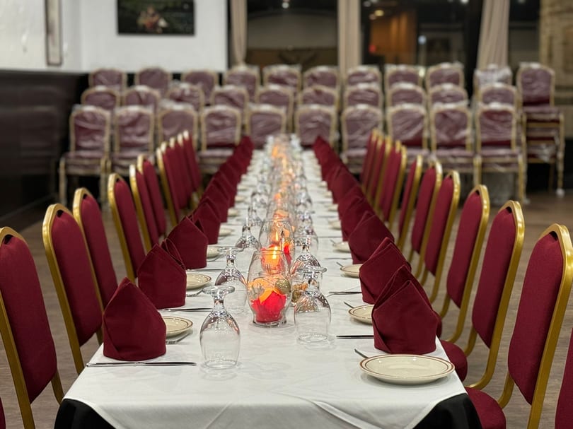 Long dining table set with white tablecloth, red napkins, plates, glasses, and candles, flanked by red chairs.