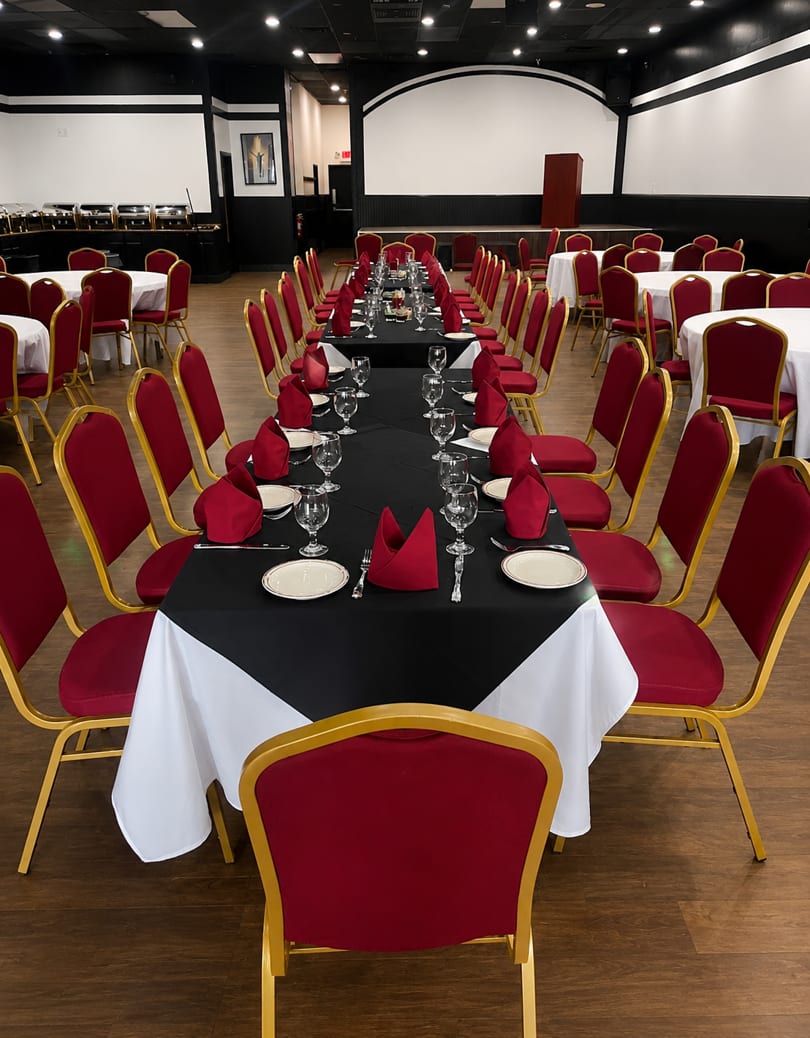 Long dining tables with black and white tablecloths, red napkins, and gold-framed red chairs in a banquet hall.